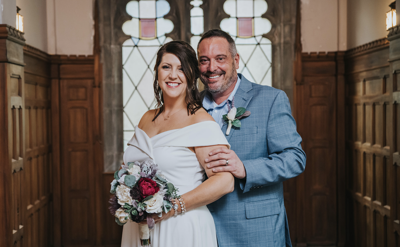 Elopement Wedding A smiling couple stands close together in a wood-paneled room with ornate windows. The woman wears an off-shoulder white dress and holds a bouquet of red, white, and pink flowers. The man, dressed in a light blue suit, stands behind her, resting his hand on her shoulder. Elopements Inc